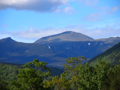 [Panoramic view of the heavily treed mountains and valley with many clouds in the sky. In the foreground is the fenced edge of the cliff.]